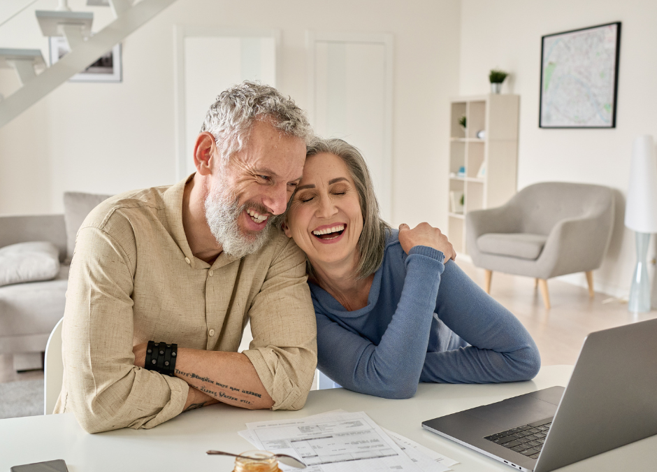 A middle-aged white couple smile with their heads together as they look at a laptop computer